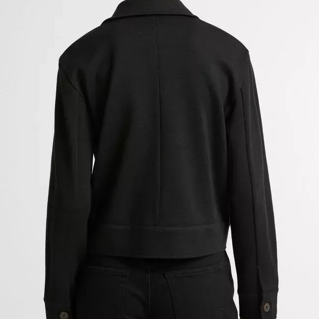 A person shown from the back wearing a black long-sleeve cropped jacket with button cuffs and black pants, photographed against a plain light gray studio background.