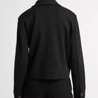A person shown from the back wearing a black long-sleeve cropped jacket with button cuffs and black pants, photographed against a plain light gray studio background.
