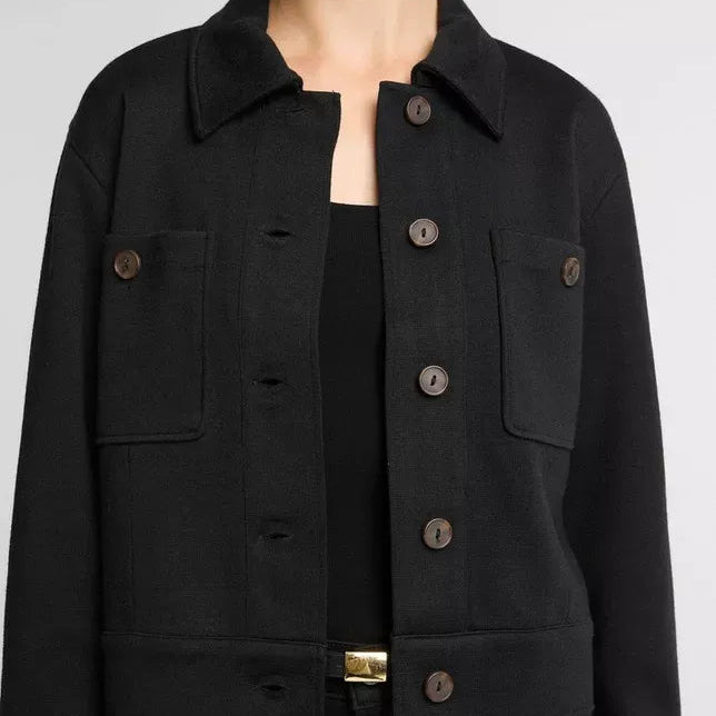 A close-up of a model wearing a black cropped jacket with a collar, button front, and flap chest pockets, paired with a black top and pants against a light gray background.