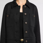 A close-up of a model wearing a black cropped jacket with a collar, button front, and flap chest pockets, paired with a black top and pants against a light gray background.