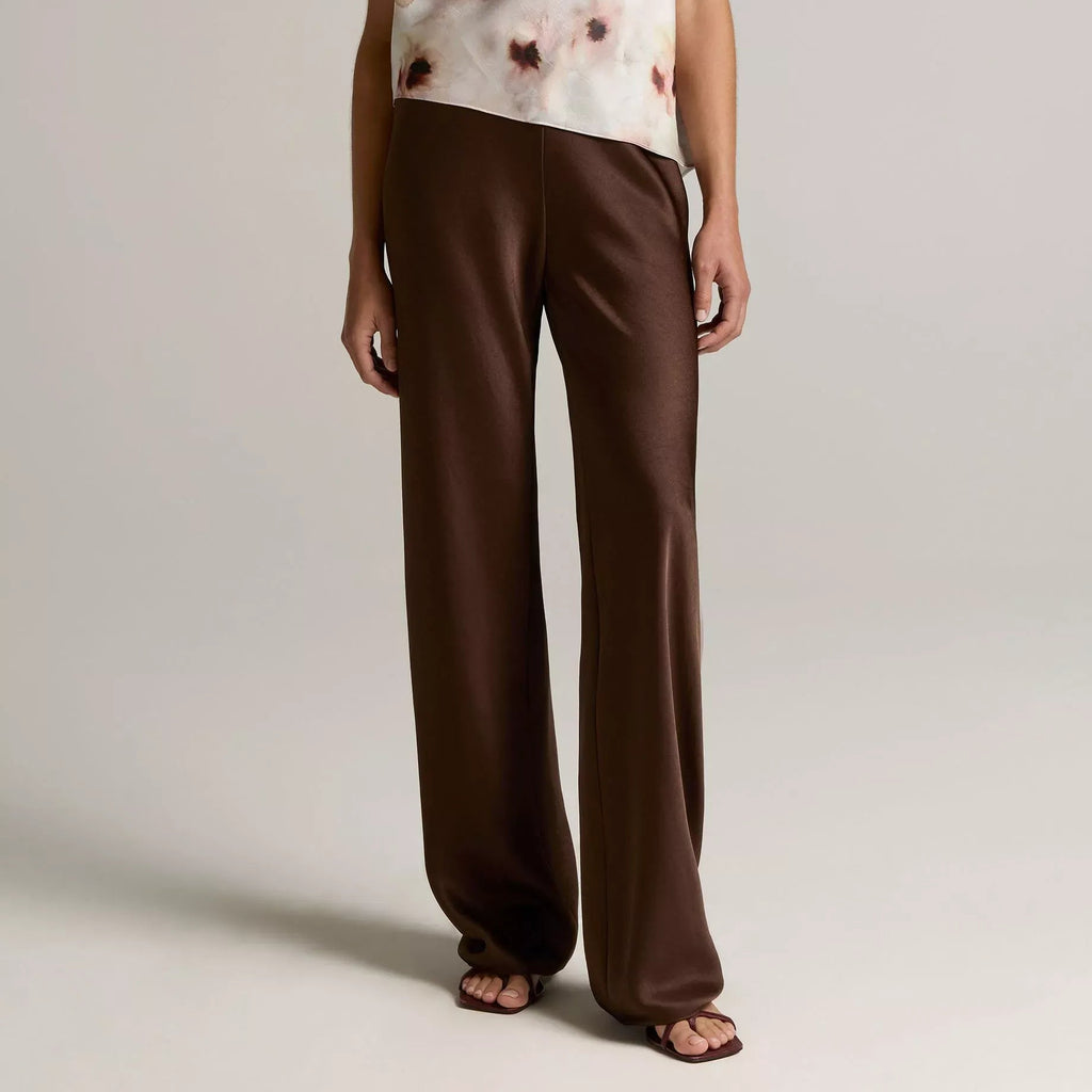 A model wearing a sleeveless draped top with a white and brown abstract print, paired with wide-leg brown trousers, against a neutral light gray studio background.