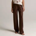 A model wearing a sleeveless draped top with a white and brown abstract print, paired with wide-leg brown trousers, against a neutral light gray studio background.