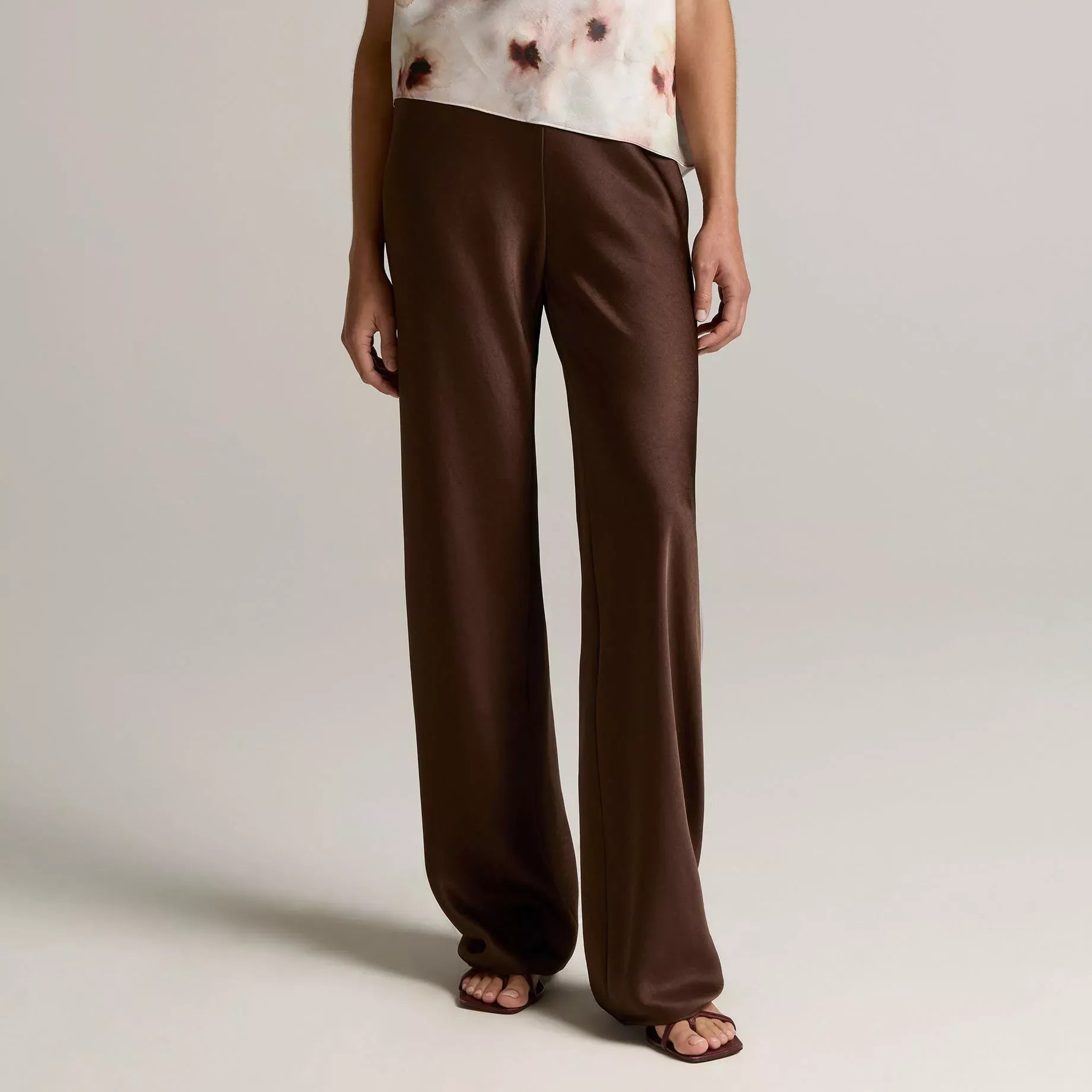 A model wearing a sleeveless draped top with a white and brown abstract print, paired with wide-leg brown trousers, against a neutral light gray studio background.