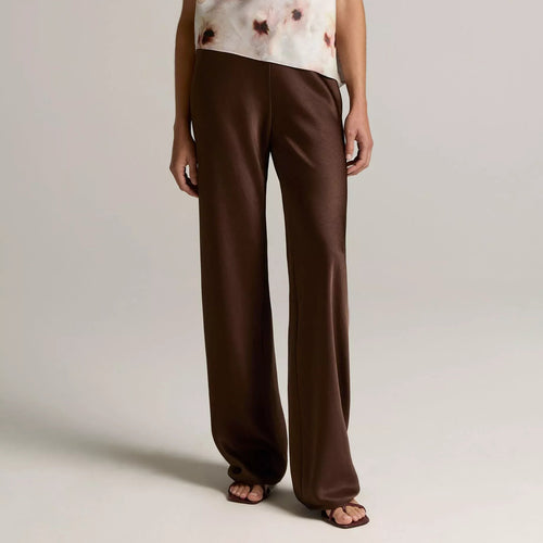 A model wearing a sleeveless draped top with a white and brown abstract print, paired with wide-leg brown trousers, against a neutral light gray studio background.