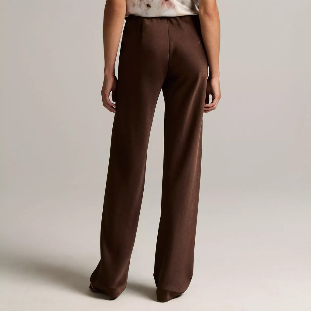 A model shown from the back wearing a sleeveless top with a white base and brown floral pattern, paired with high-waisted brown wide-leg trousers, against a neutral light gray studio background.