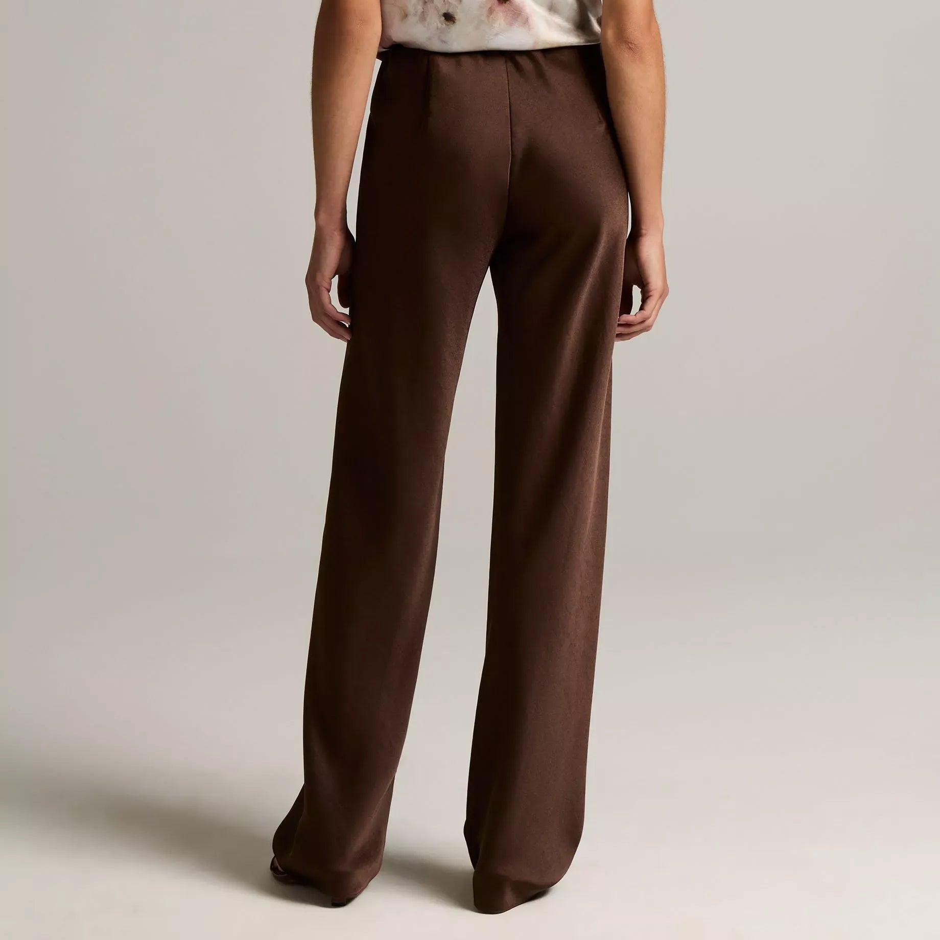 A model shown from the back wearing a sleeveless top with a white base and brown floral pattern, paired with high-waisted brown wide-leg trousers, against a neutral light gray studio background.