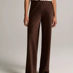 A model wearing high-waisted, wide-leg brown trousers paired with a sleeveless floral top and flat sandals, shown from the chest down against a neutral studio background.