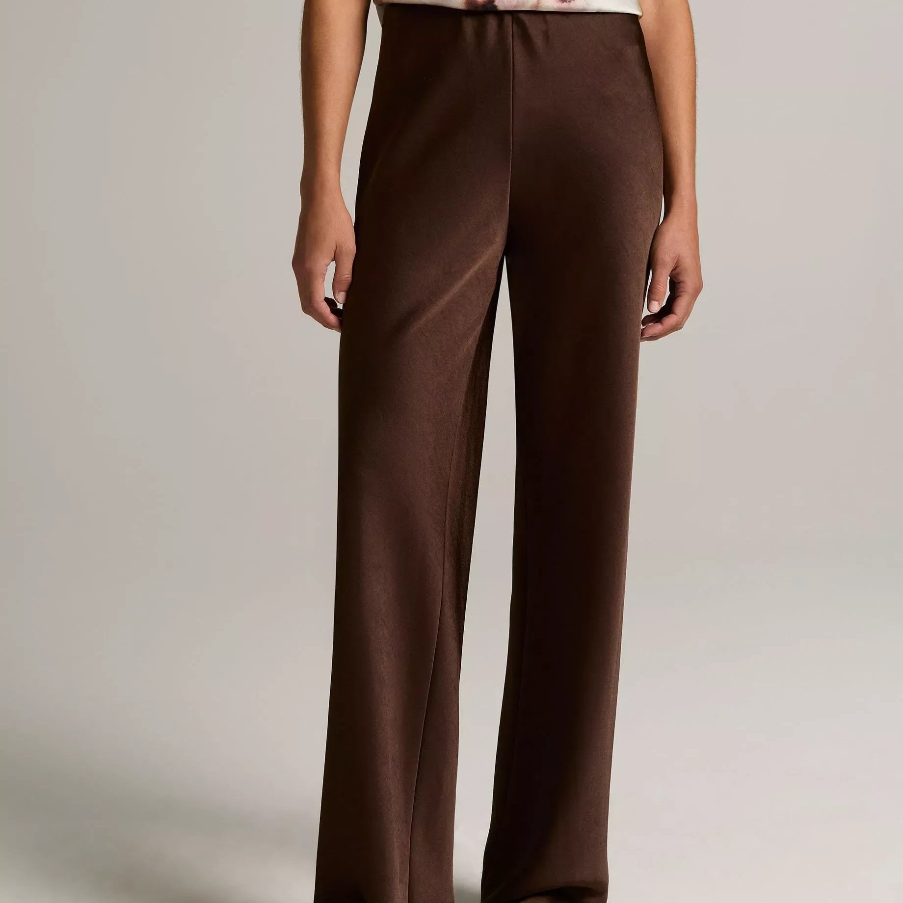 A model wearing high-waisted, wide-leg brown trousers paired with a sleeveless floral top and flat sandals, shown from the chest down against a neutral studio background.