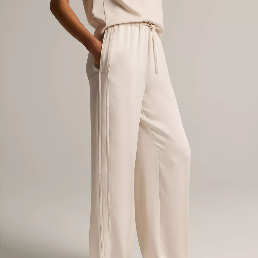 A model wearing a pair of wide-leg, cream-colored trousers with a drawstring waist, paired with a matching sleeveless top and flat sandals, set against a plain light gray studio background.
