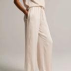 A model wearing a pair of wide-leg, cream-colored trousers with a drawstring waist, paired with a matching sleeveless top and flat sandals, set against a plain light gray studio background.