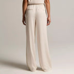 A model shown from the back wearing a sleeveless cream jumpsuit with wide-leg trousers and a high-waisted design, set against a plain light gray studio background.