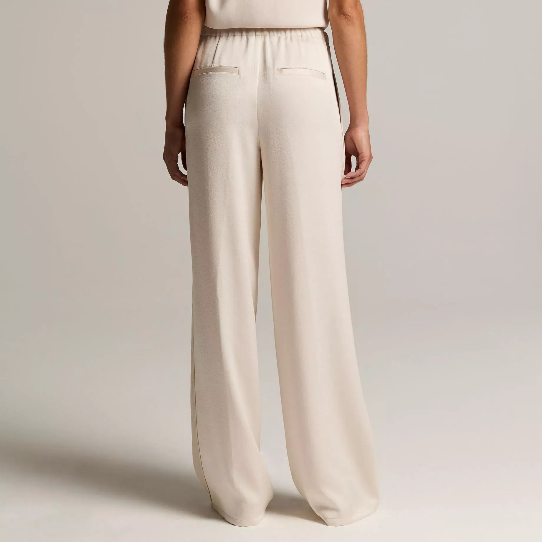 A model shown from the back wearing a sleeveless cream jumpsuit with wide-leg trousers and a high-waisted design, set against a plain light gray studio background.