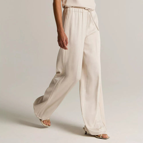 A model wearing a light cream sleeveless top and matching wide-leg trousers with a tie belt, styled with beige heeled sandals against a neutral studio background.