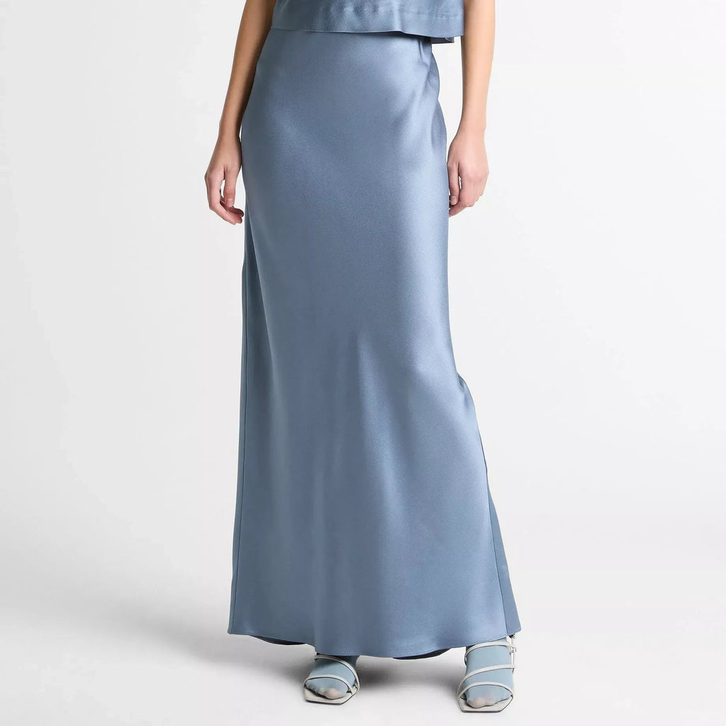 A model wearing a two-piece satin outfit in a dusty blue color, featuring a sleeveless mock-neck top and a matching long skirt, paired with light blue strappy sandals against a plain white studio background.