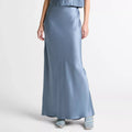 A model wearing a two-piece satin outfit in a dusty blue color, featuring a sleeveless mock-neck top and a matching long skirt, paired with light blue strappy sandals against a plain white studio background.