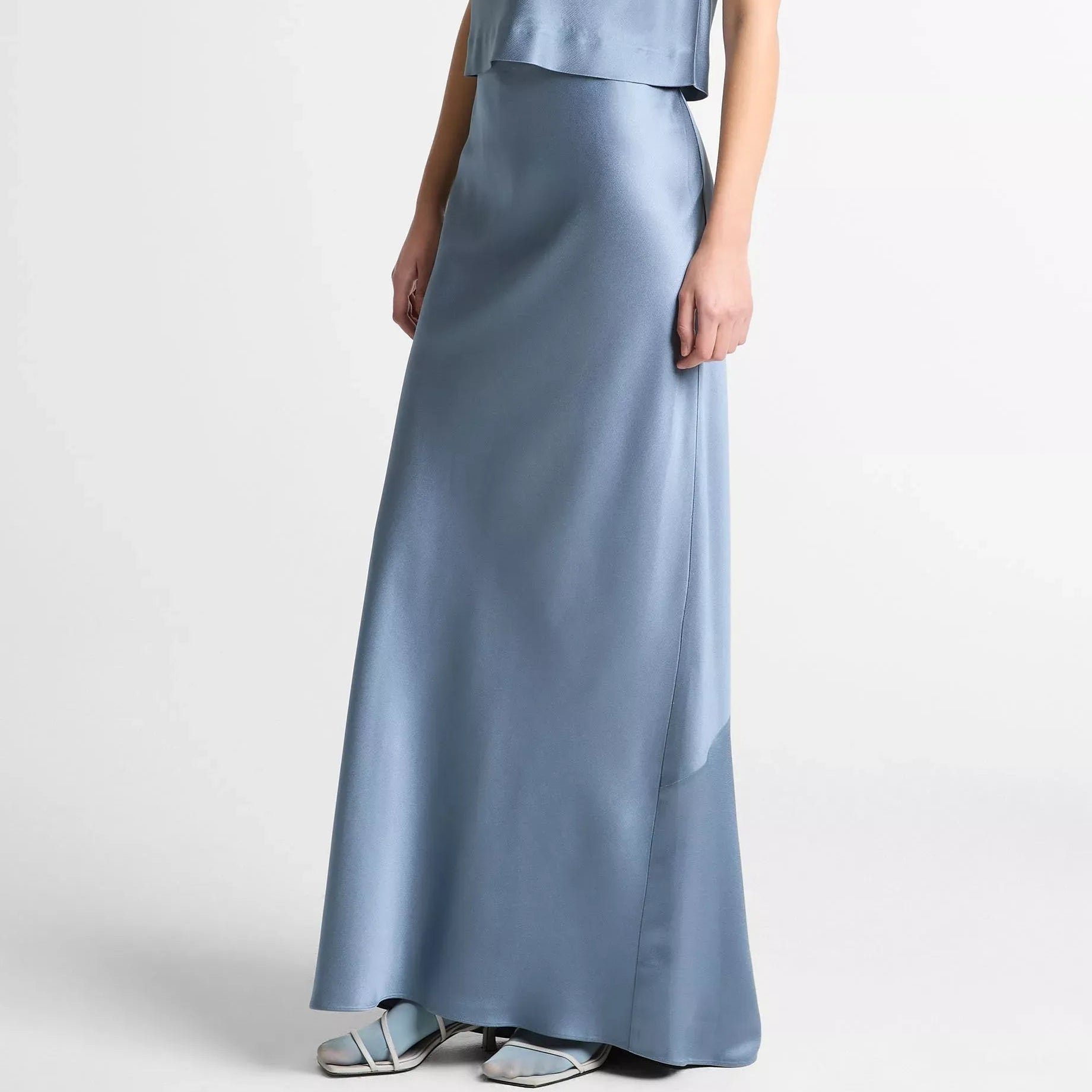 A model wearing a light blue satin two-piece set consisting of a sleeveless draped cowl-neck top and a matching long skirt, paired with silver flat sandals against a light gray studio background.