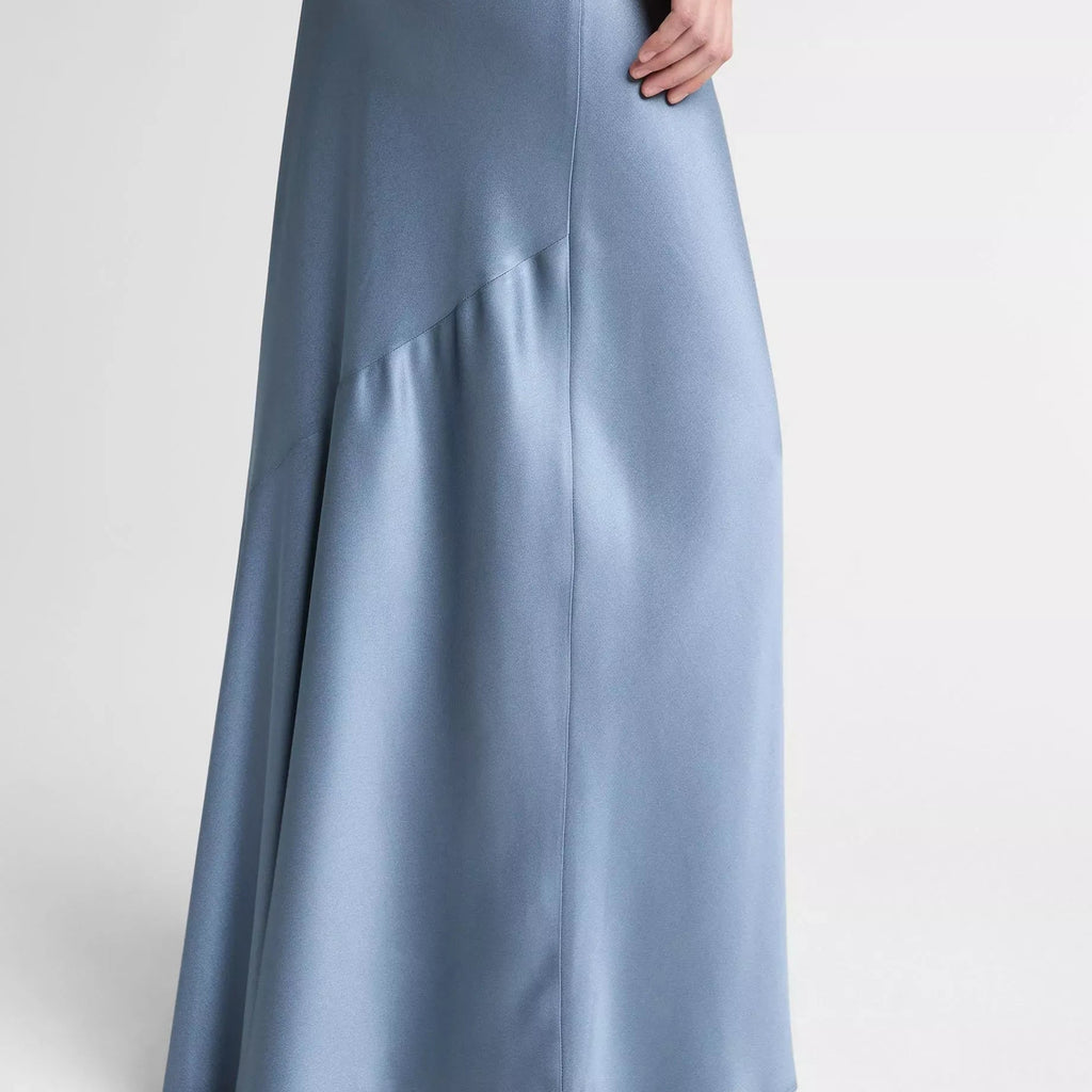A model wearing a floor-length, light blue satin maxi skirt with a draped asymmetrical hem and silver pointed-toe heeled sandals, shown from the waist down against a light gray studio background.