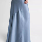 A model wearing a floor-length, light blue satin maxi skirt with a draped asymmetrical hem and silver pointed-toe heeled sandals, shown from the waist down against a light gray studio background.
