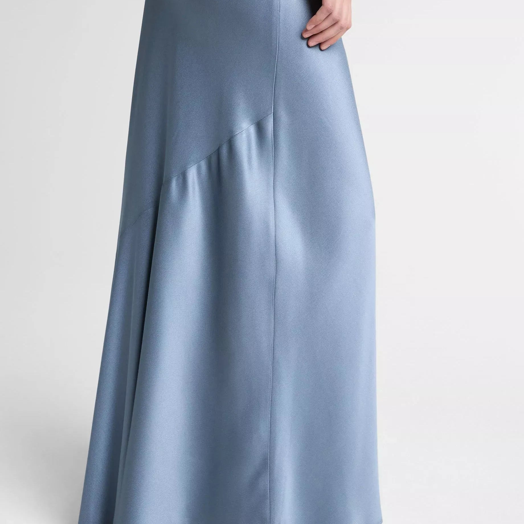 A model wearing a floor-length, light blue satin maxi skirt with a draped asymmetrical hem and silver pointed-toe heeled sandals, shown from the waist down against a light gray studio background.