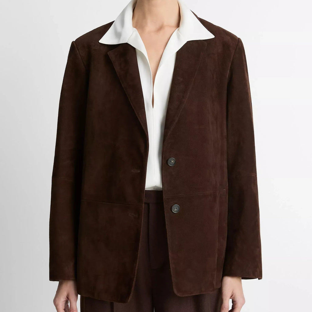 A model wearing a dark brown suede blazer with a contrasting white collar, paired with a white shirt and matching dark brown trousers, shot against a light gray studio background.