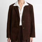 A model wearing a dark brown suede blazer with a contrasting white collar, paired with a white shirt and matching dark brown trousers, shot against a light gray studio background.