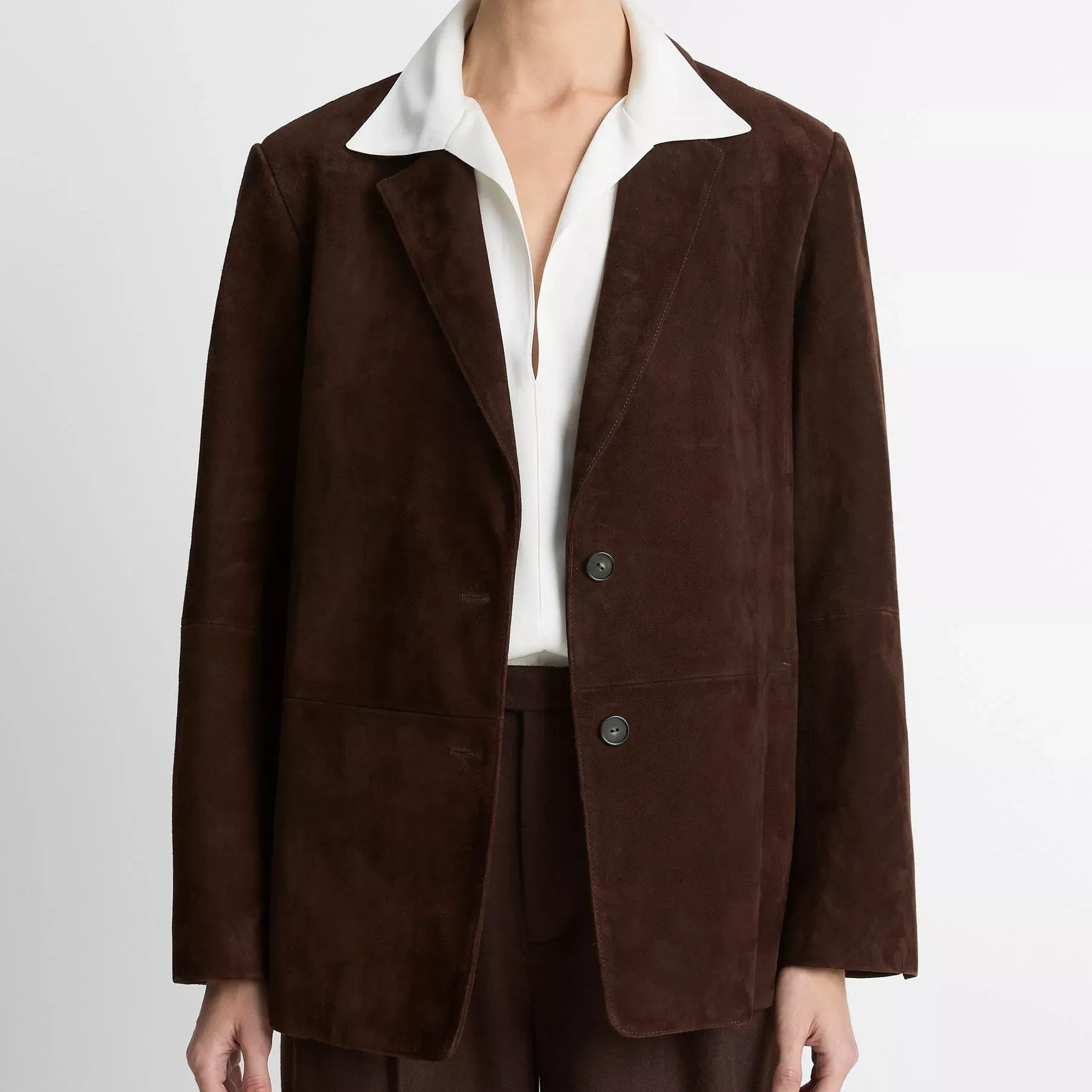 A model wearing a dark brown suede blazer with a contrasting white collar, paired with a white shirt and matching dark brown trousers, shot against a light gray studio background.