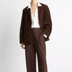 A model wearing a dark brown suede jacket and matching wide-leg trousers, styled with a white collared shirt and beige pointed-toe flats, against a plain light gray studio background.