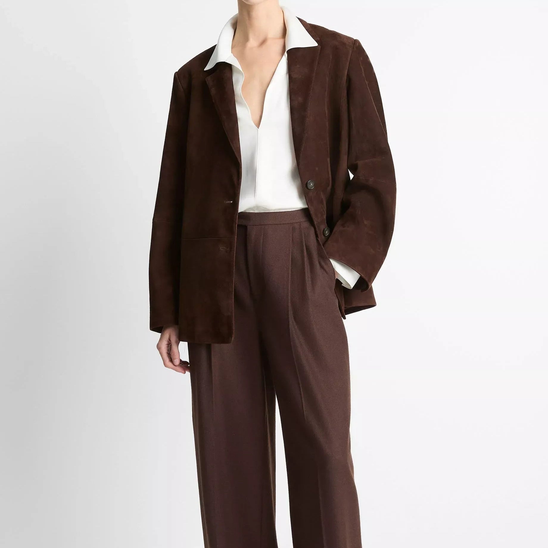 A model wearing a dark brown suede jacket and matching wide-leg trousers, styled with a white collared shirt and beige pointed-toe flats, against a plain light gray studio background.