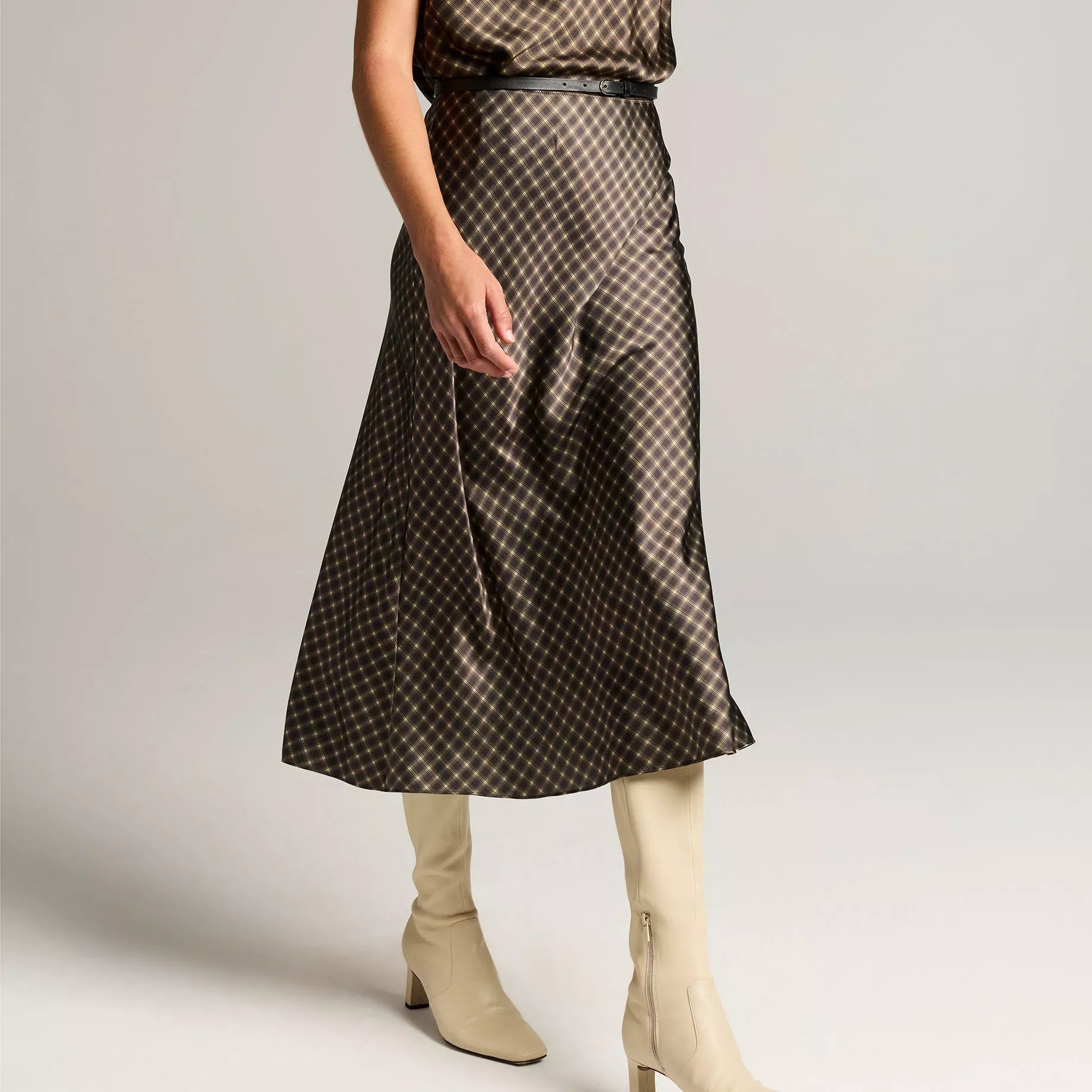 A model wearing a brown and black patterned midi skirt with a matching cowl neck top, paired with beige heeled ankle boots, against a light gray studio background.