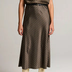 A model wearing a brown and beige patterned midi skirt with a thin black belt, paired with beige ankle boots, against a light gray studio background.