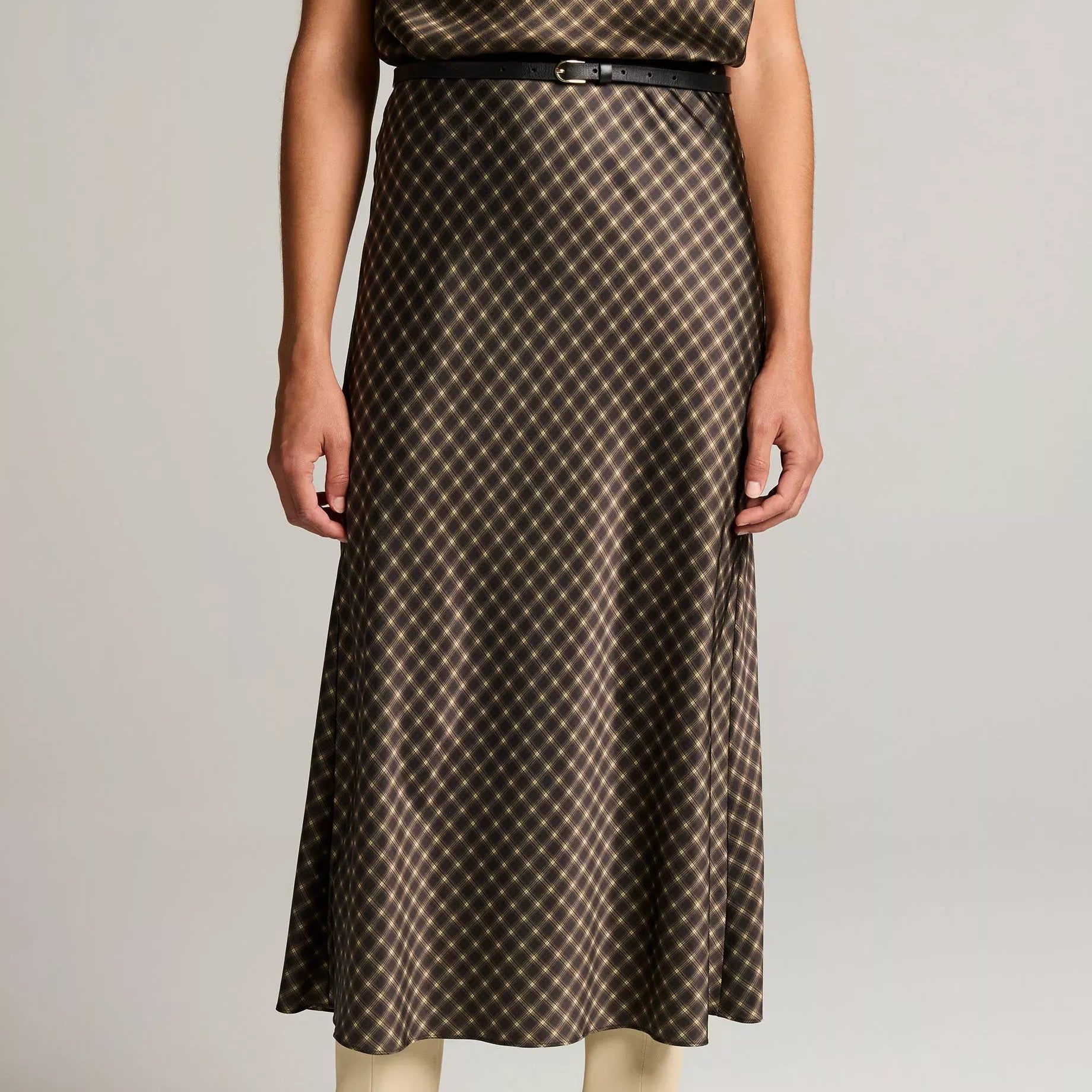 A model wearing a brown and beige patterned midi skirt with a thin black belt, paired with beige ankle boots, against a light gray studio background.