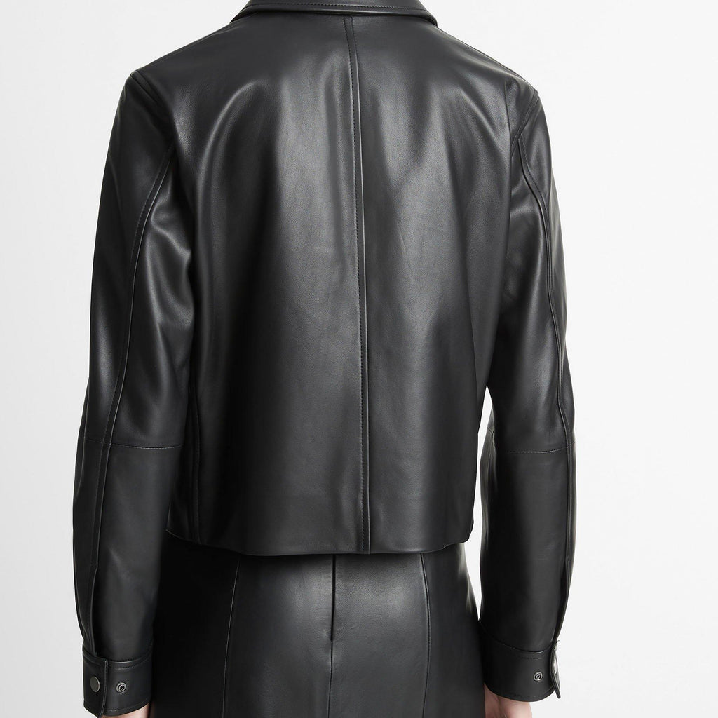 Rear view of a model wearing a black leather cropped jacket and matching black leather skirt against a plain light gray studio background.