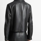 Rear view of a model wearing a black leather cropped jacket and matching black leather skirt against a plain light gray studio background.