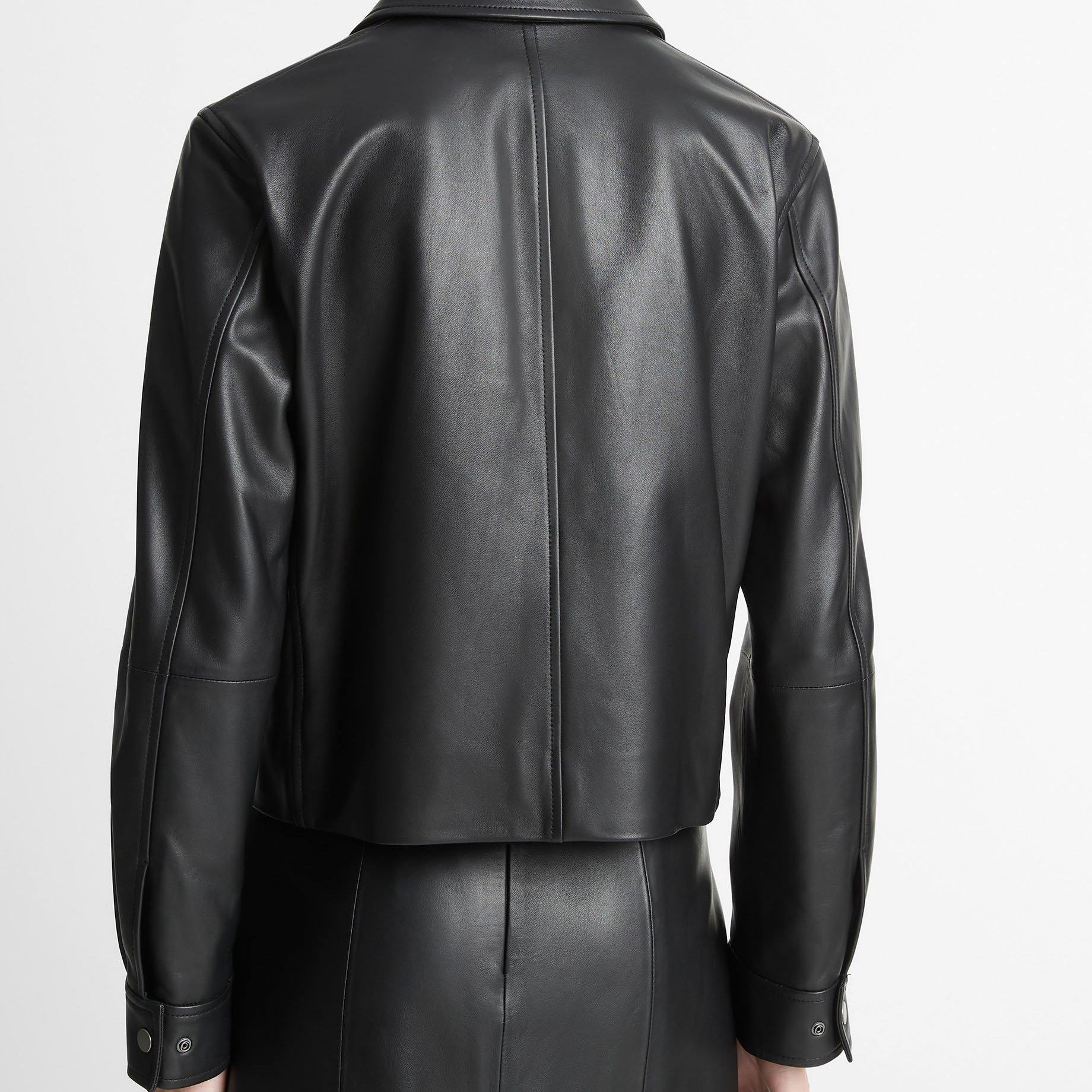 Rear view of a model wearing a black leather cropped jacket and matching black leather skirt against a plain light gray studio background.