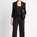 A model wearing a black tailored pantsuit consisting of a single-breasted blazer with three-quarter sleeves, a scoop-neck top, and wide-leg trousers, styled with black open-toe heels against a plain white studio background.