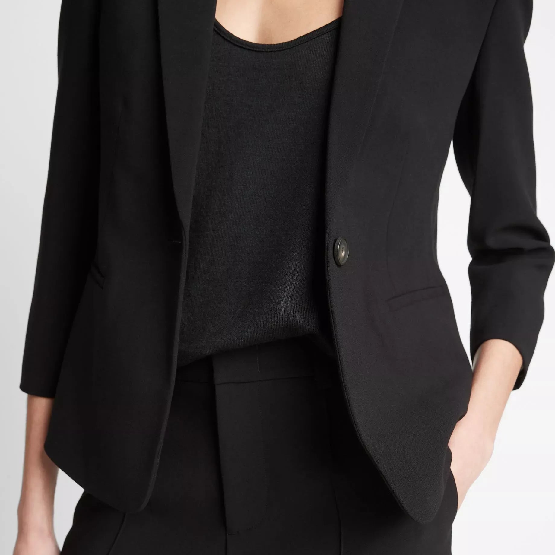 A close-up of a model wearing a black tailored blazer with three-quarter sleeves, paired with a black top and black trousers, against a light gray studio background.