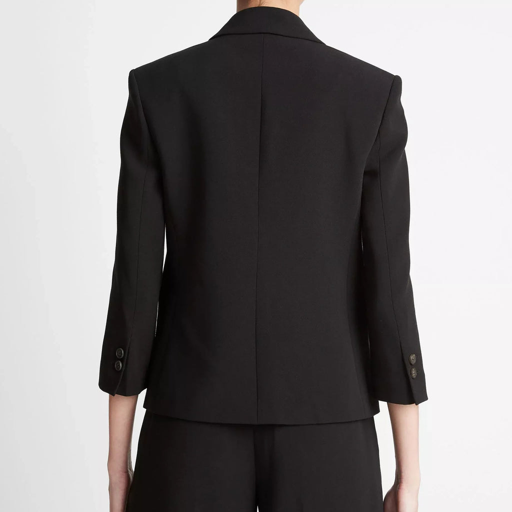 Rear view of a model wearing a black tailored blazer with three-quarter sleeves and matching black trousers against a plain light gray studio background.