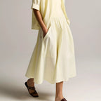 A woman modeling a light cream-colored linen dress with a matching short-sleeve cardigan, styled with dark brown leather slide sandals against a neutral studio background.