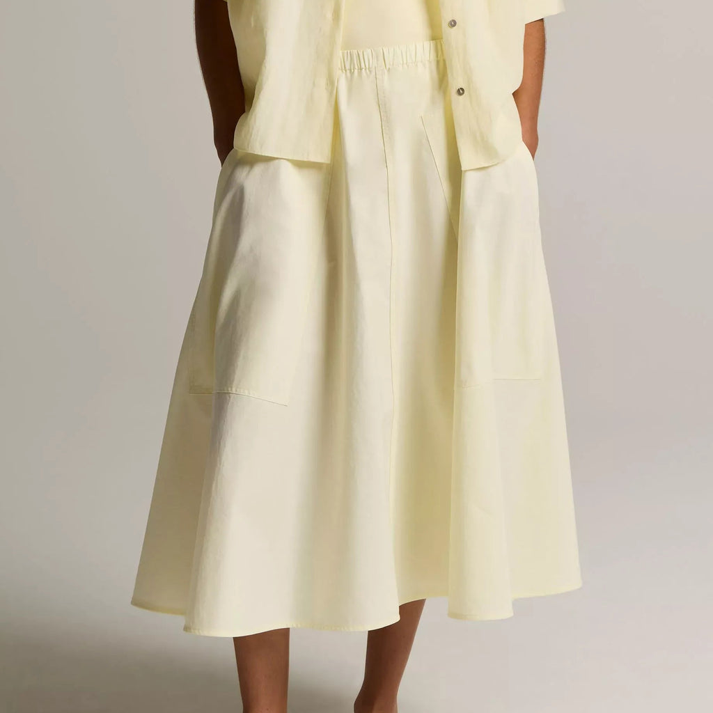 A model wearing a light yellow linen midi dress with a matching short-sleeve button-up shirt and dark brown leather slide sandals, shown from the waist down against a neutral studio background.