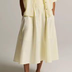 A model wearing a light yellow linen midi dress with a matching short-sleeve button-up shirt and dark brown leather slide sandals, shown from the waist down against a neutral studio background.