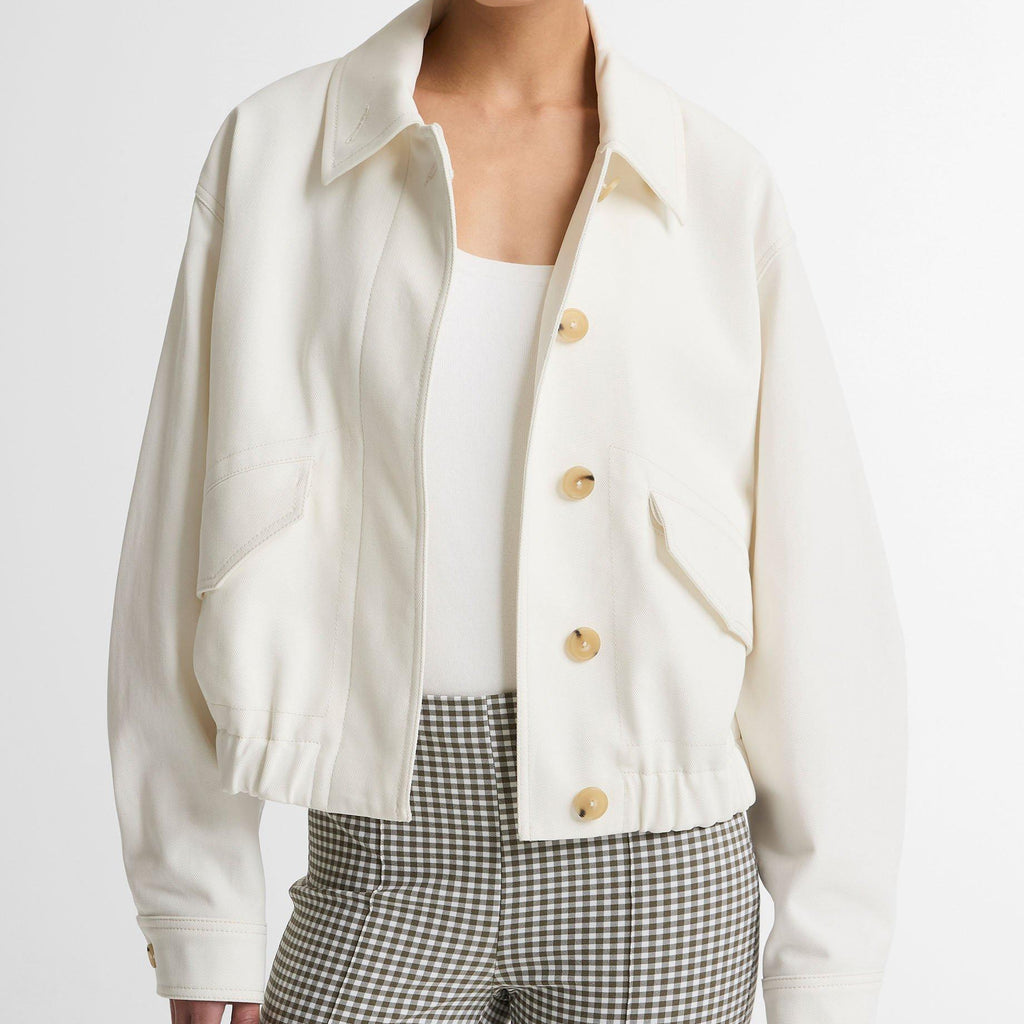 A model wearing a cropped, off-white button-up jacket with gold buttons, paired with a white top and green-and-white checkered trousers, against a plain light gray background.