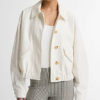 A model wearing a cropped, off-white button-up jacket with gold buttons, paired with a white top and green-and-white checkered trousers, against a plain light gray background.
