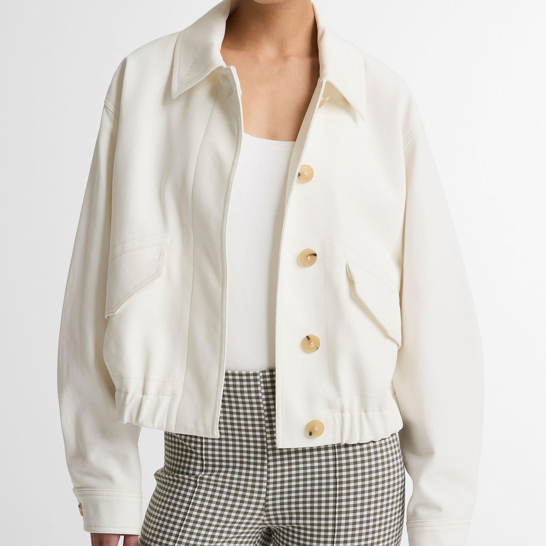 A model wearing a cropped, off-white button-up jacket with gold buttons, paired with a white top and green-and-white checkered trousers, against a plain light gray background.