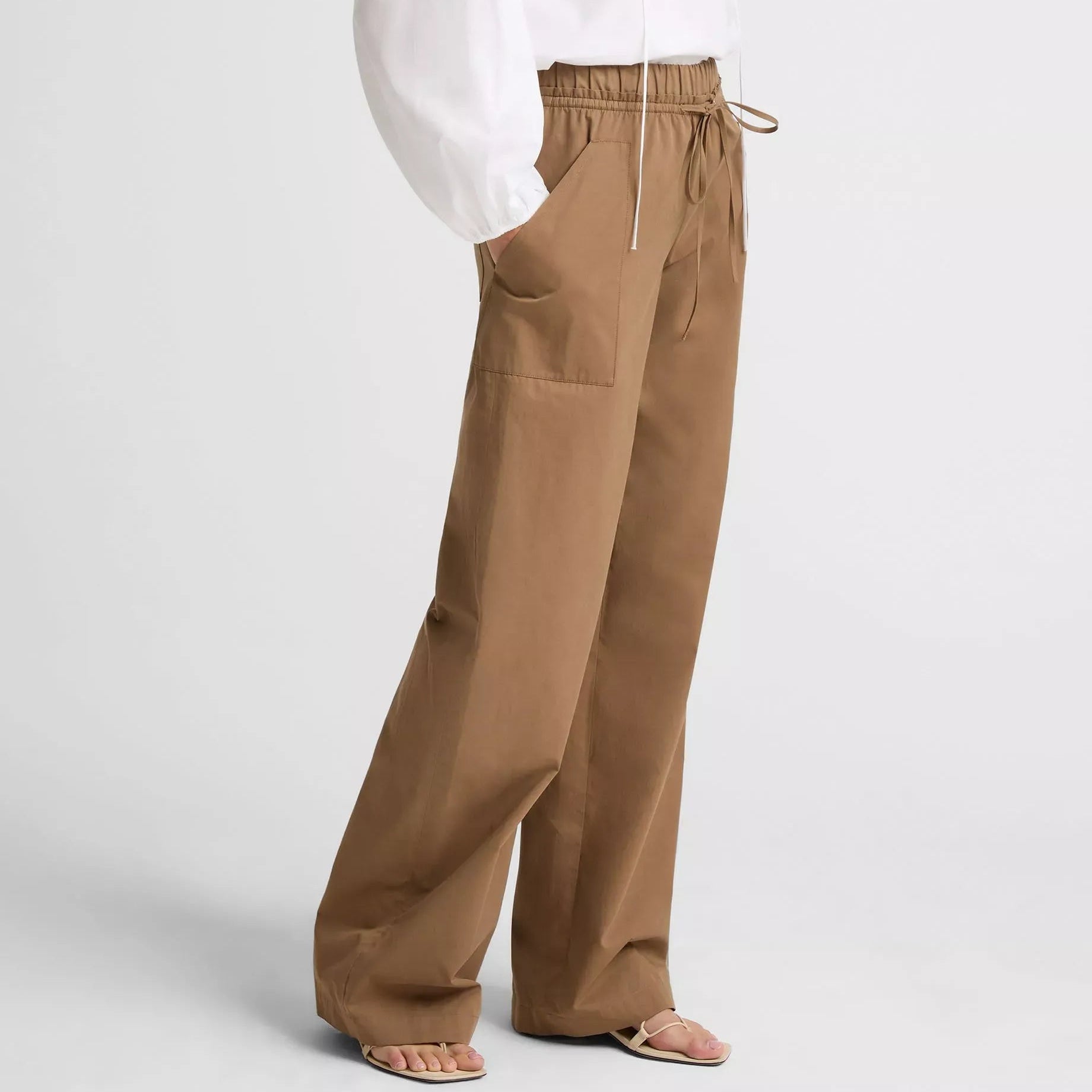 A model wearing a light beige sleeveless top and matching wide-leg trousers with a tie waist, styled with beige heeled sandals against a neutral studio background.