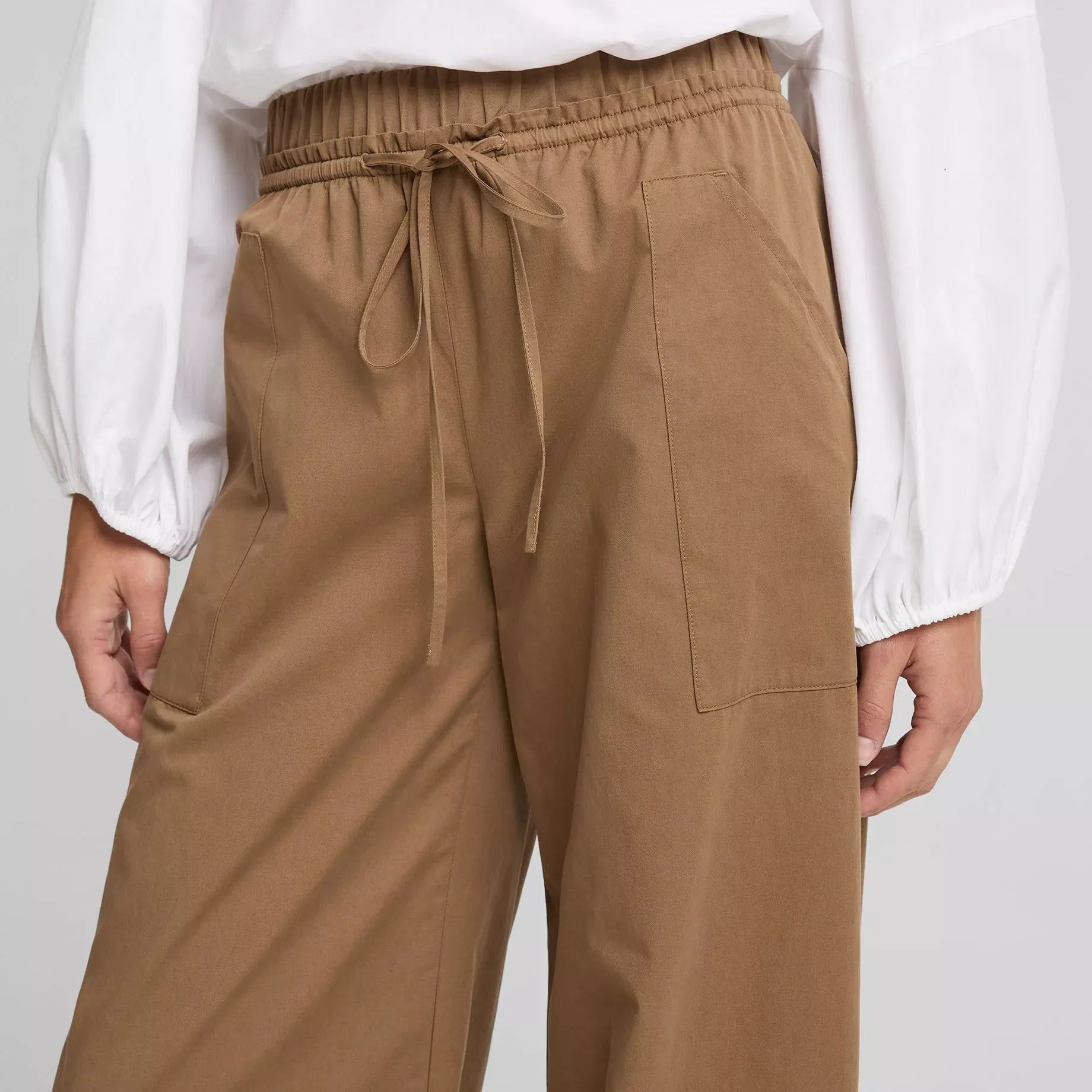 A person wearing brown wide-leg drawstring pants and a white long-sleeve blouse, shown from the chest down against a light gray studio background.